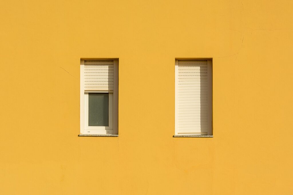 A yellow wall featuring two closed windows with roller shutters, showcasing a minimalist design.